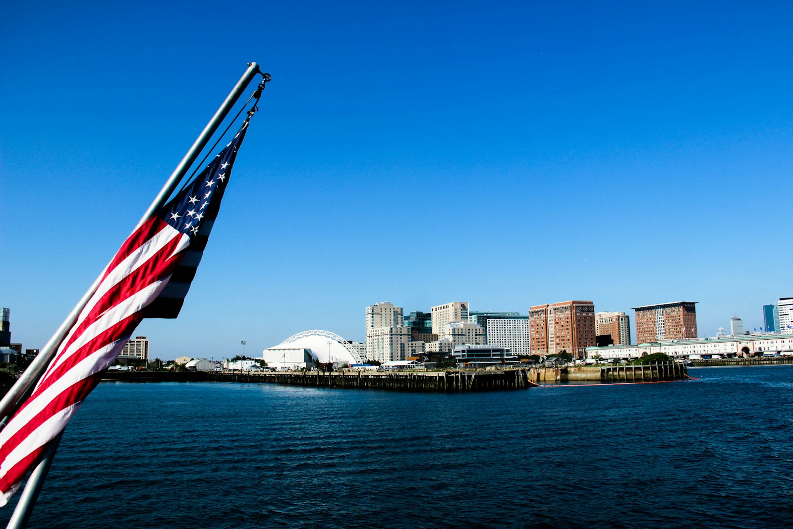 An American flag waving above a harbor with a city skyline in the background